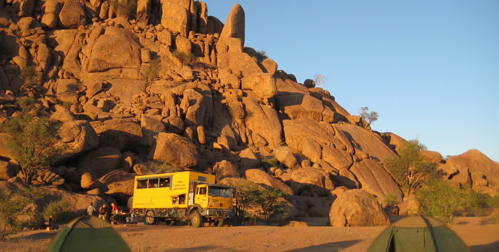 A yellow bus against desert cliffs with green tents dotted in the foreground