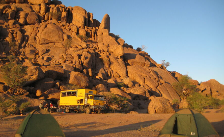 A yellow bus against desert cliffs with green tents dotted in the foreground