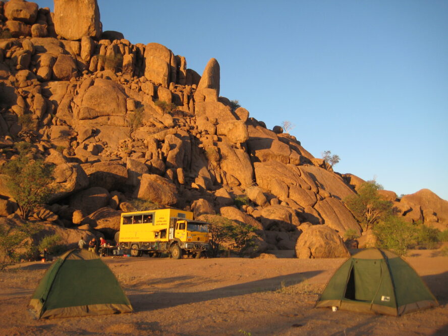 A yellow bus against desert cliffs with green tents dotted in the foreground