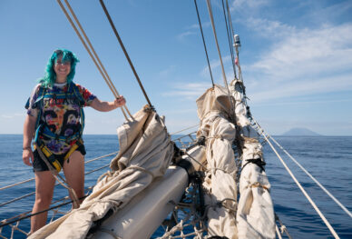 A blue haired woman stands at the front of the ship - volcano in the background