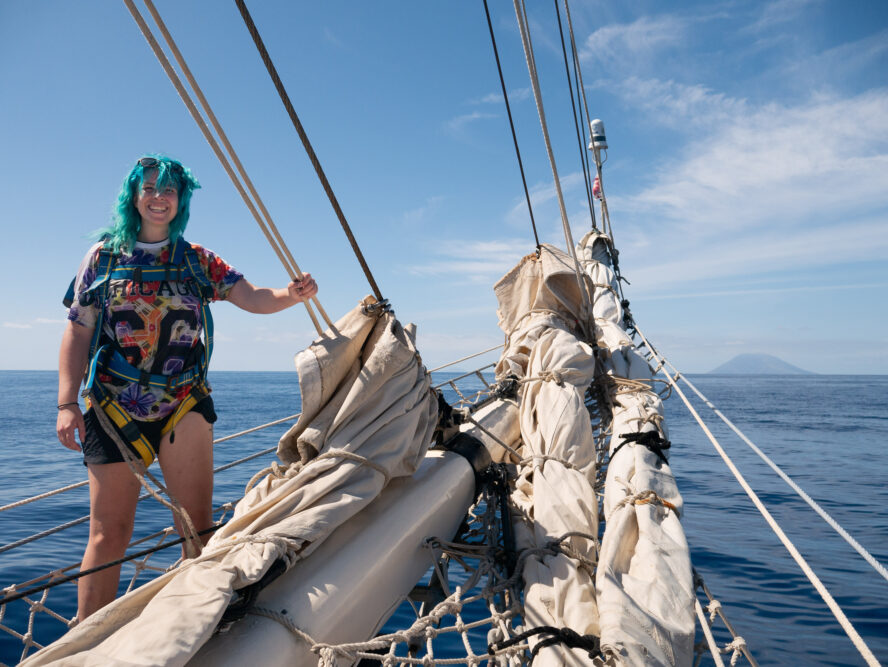 A blue haired woman stands at the front of the ship - volcano in the background