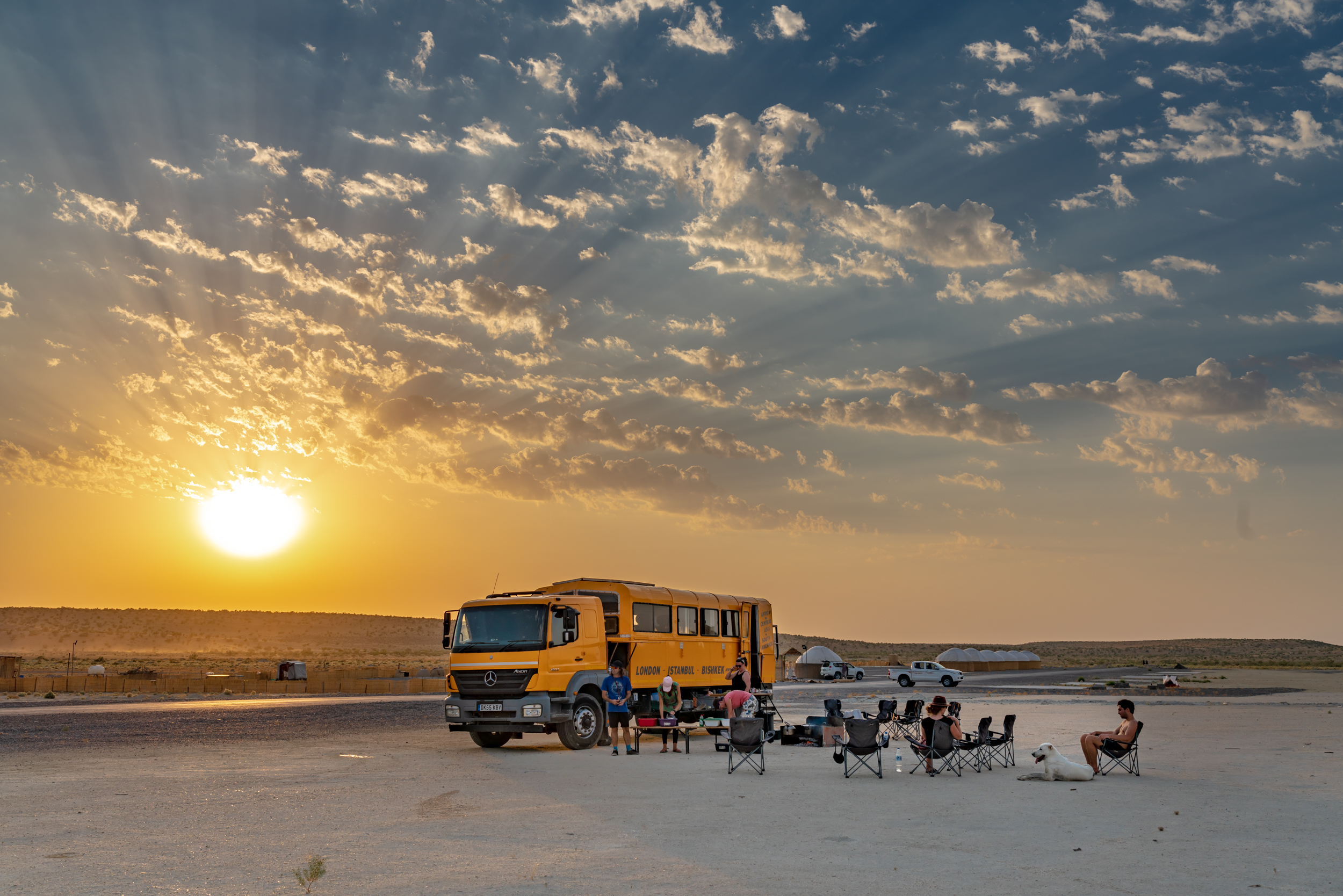 A sun sinking creating ripples in the sky, a large yellow truck seems small in the frame with a camp set out around it