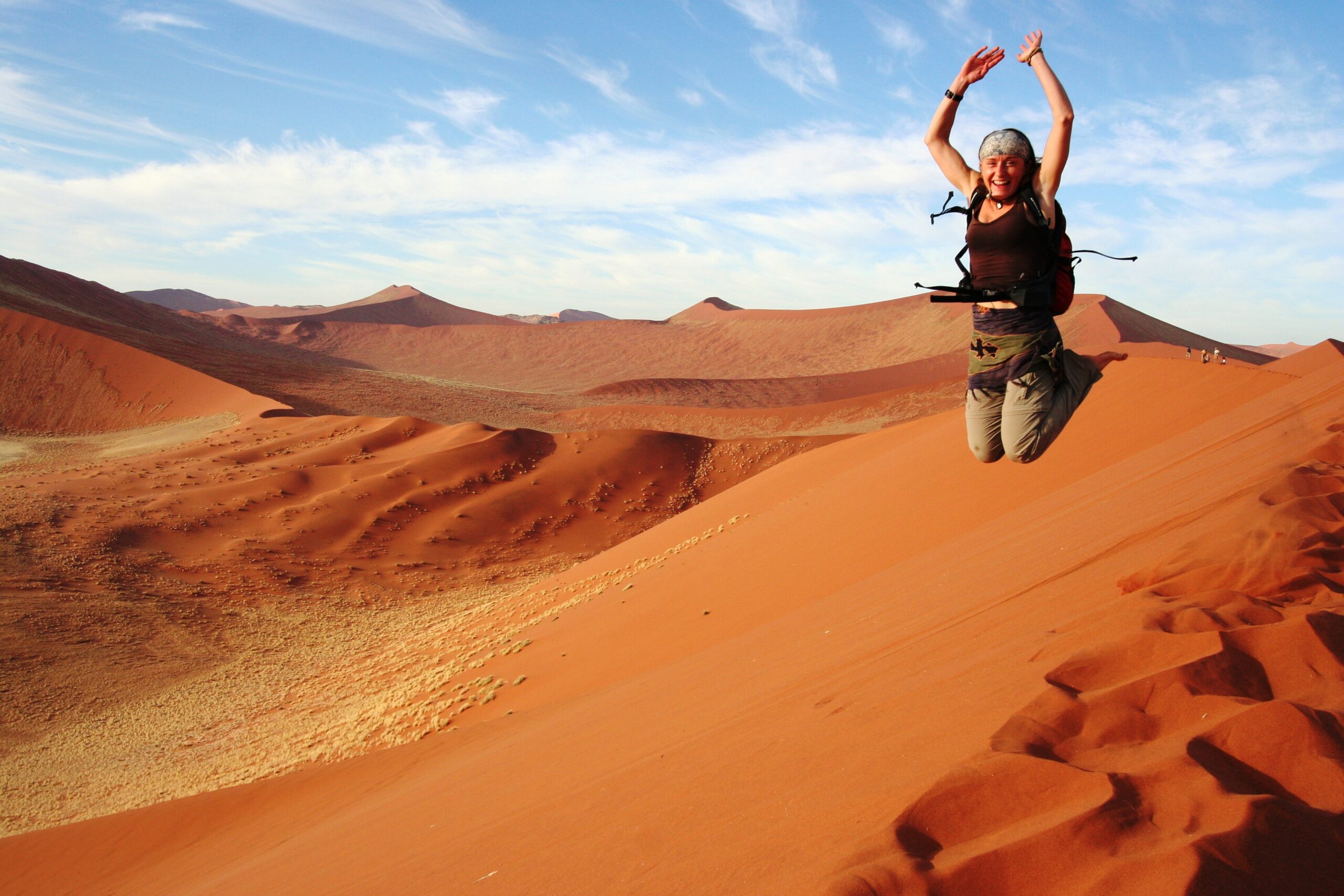 A woman jumps into the air with a bright orange sand dune snaking behind her